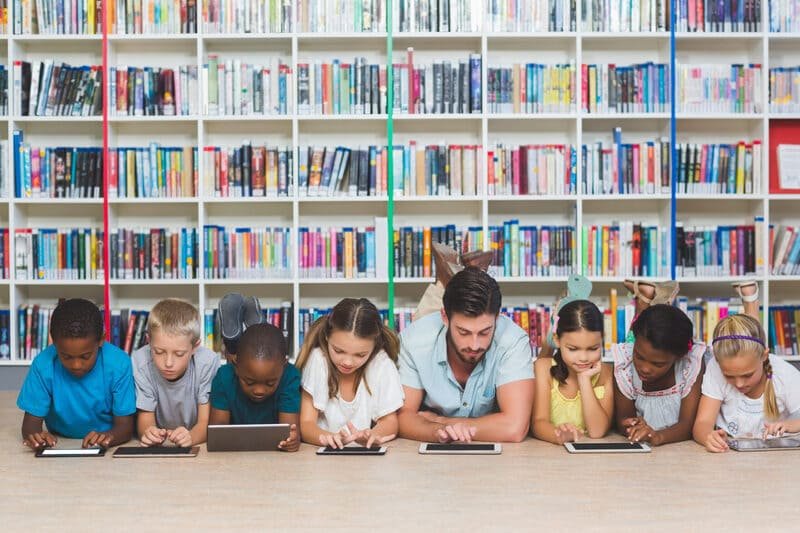 Teacher and young students engaged with iPads in a library, enhancing learning through technology provided by iPadify, accessible for educational institutions at discounted rates.